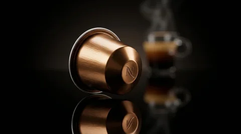 Uma cápsula de café metálica cor de cobre deitada de lado sobre uma superfície preta altamente reflexiva, exibindo um reflexo nítido. A cápsula possui uma textura escovada elegante e a letra "N" em relevo na sua base. Ao fundo, levemente desfocada, há uma xícara de vidro transparente com café expresso fumegante, compondo um ambiente escuro e sofisticado com iluminação dramática.